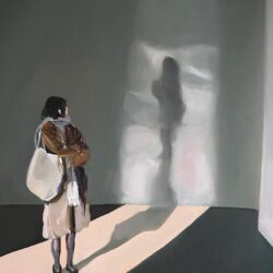 A woman stands in an open room and looks back over her left shoulder. In the back is the shadow of the woman against a wall.