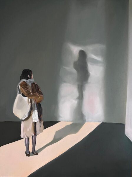 A woman stands in an open room and looks back over her left shoulder. In the back is the shadow of the woman against a wall.