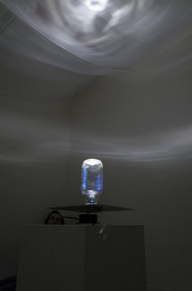 Installation of a big rounded shaped glass tube with a light inside, illuminating the ceiling from the top part of the glass tube from a table, and behind the stand where the emitting light sculpture si placed, is visible the face of a person in the room corner