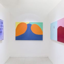 Three paintings installed, one on each wall from left to right the paintings are purple, black, the middle painting is blue with orange round shapes and a yellow and green background, the left has a painting that is half light blue, and half pink with a big red round shape