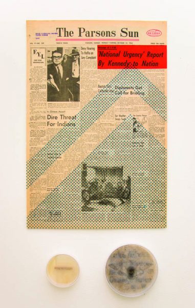 A page of The Parson Sun with the title "National Urgency" Report by Kennedy to Nation. The news article has text and two black and white images with a man. Under the newspaper page are two rounded containers, one with a yellow organic material and the other containing a preserved insect