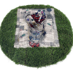 Sculpture of a folded newspaper with attached images encapsulated in epoxy on a green turf base