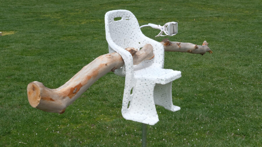 A mixed-media sculpture with a white mosaic tile covered chair and a large light colored branch inserted through the chair