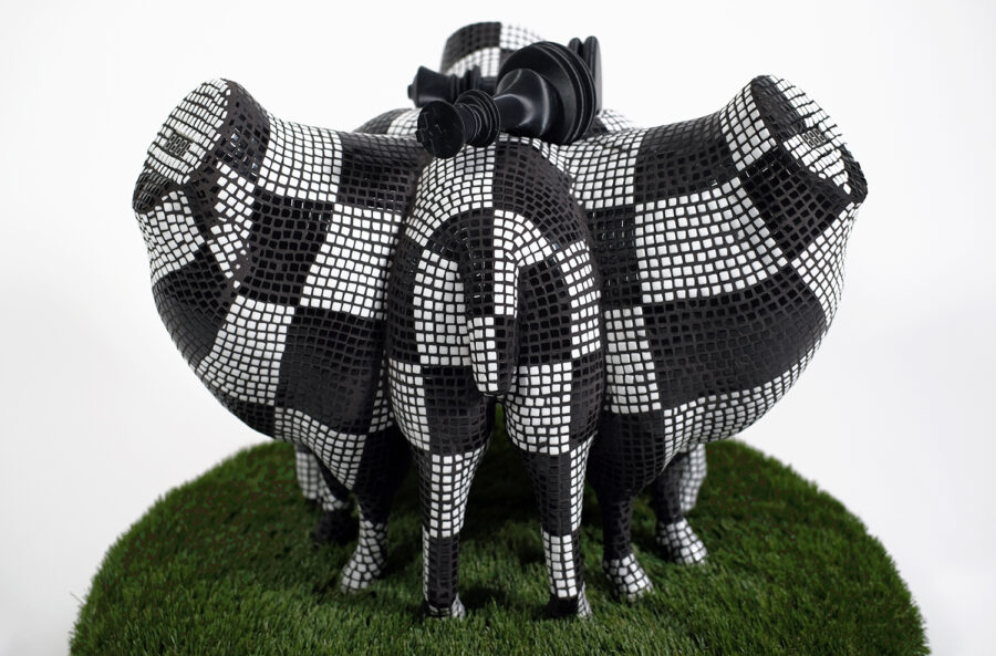 A sculpture featuring the partial bodies of multiple sheep, headless and intersecting, covered with black and white tile in a grid patter. The neck stumps of the sheep feature an embedded four-character alphanumeric 7-segment LED display. The sculpture stands on a mound of artificial grass.