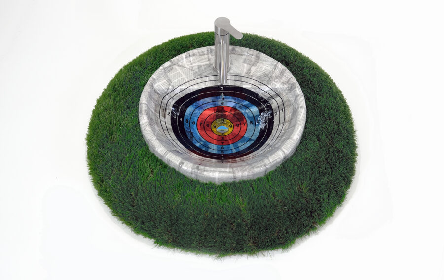 A round sink bowl decoupaged with a radial target images, with a chrome faucet, set into a mount of artificial grass.