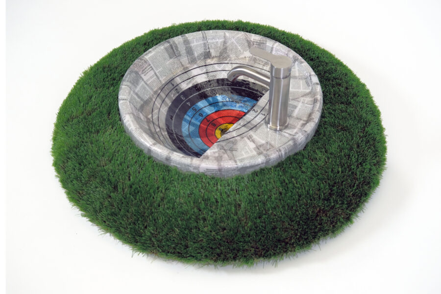 Sculpture featuring a target made from newspaper in the form of a sink basin situated on a round ring of artificial grass, there is a faucet attached to the target