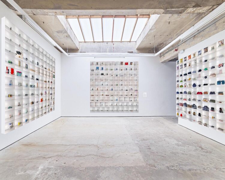 An installation view of three sets of gridded photographs depicting miscellaneous objects. The floor is concrete, the walls are white, and there is a skylight in the ceiling.