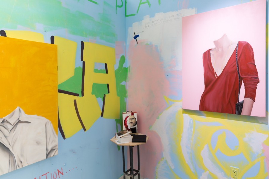 Installation view of paintings and small objects on a small table, representing portraits without heads with persons wearing a white shirt on an orange background on the left, and on the right a portrait of a person wearing a red dress with a deep cleavage and a black purse on a pink backdrop. The paintings are installed on a vivid colored wall with graffiti and some words
