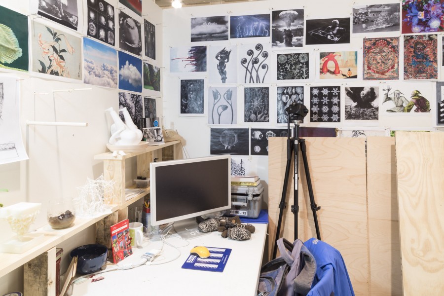 Working desk, an array of prints on the wall, a computer monitor, a tripod, and many objects on the desk.