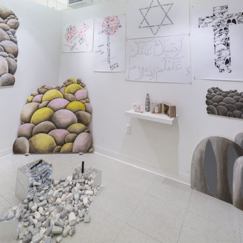 Gallery view, on the floor there are a bunch of white and gray rock sculptures installed in a pile, there are paintings of rocks on the left, middle, and right of the installation, there are also other drawings with text, a pentagram, and a cross on the wall as well as a small wall shelf with various small rock sculptures