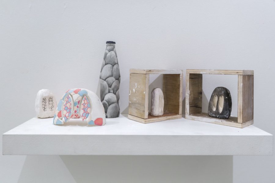 Various sculptures installed on a white wall shelf, from left to right there is a white rock sculpture, a white, blue, red sculpture, a tall vase like gray sculpture that has rocks painted on it, two rock sculptures one white and one black installed in wooden frames on the right