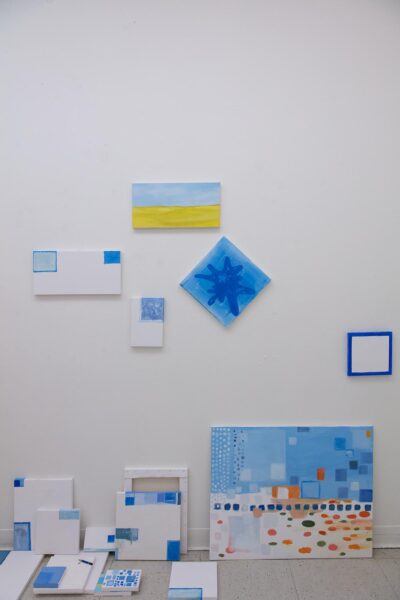 Several small paintings by Zihao Ren in blue, orange and yellow hanging on the studio wall and stacked on the floor.