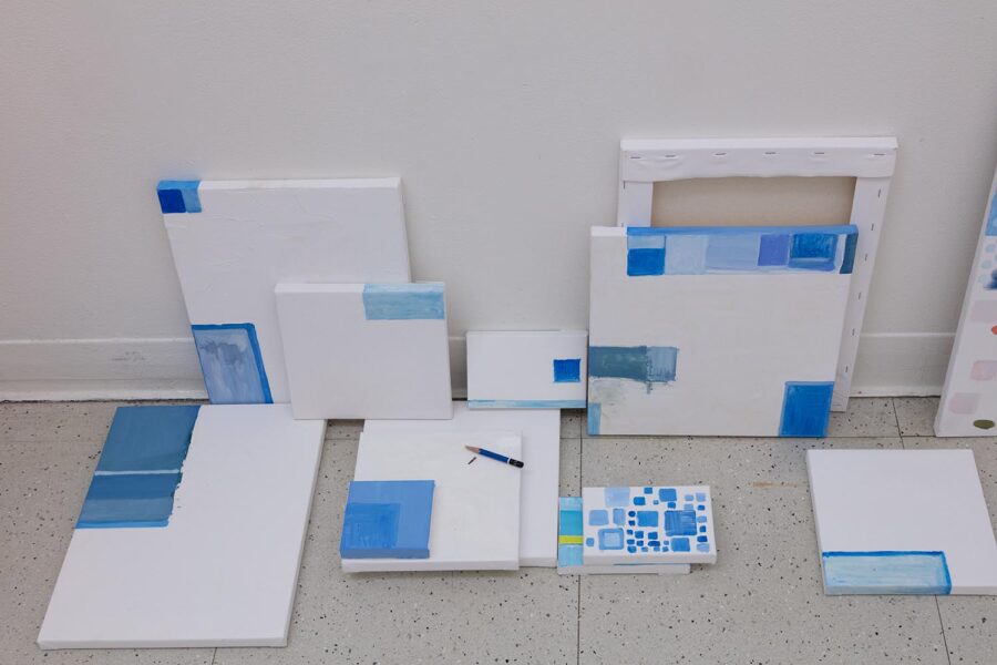 Several small paintings by Zihao Ren consisting of blue squares painted on white backgrounds sitting on the floor and leaning against the wall.