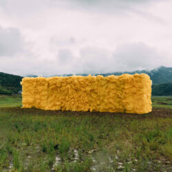 Video still of a large rectangular yellow sculpture of wood and plastic bags installed outdoors