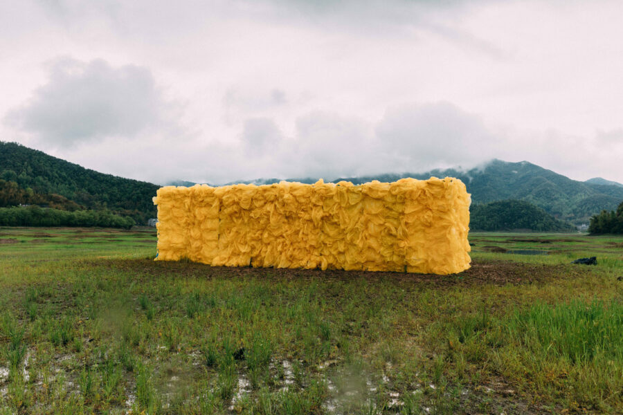 Video still of a large rectangular yellow sculpture of wood and plastic bags installed outdoors