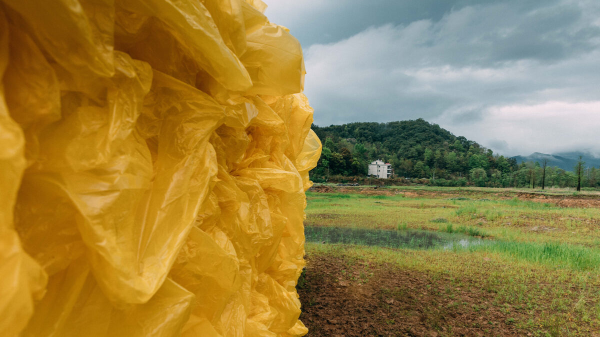 Close-up video still of a large rectangular yellow sculpture of wood and plastic bags installed outdoors with a house