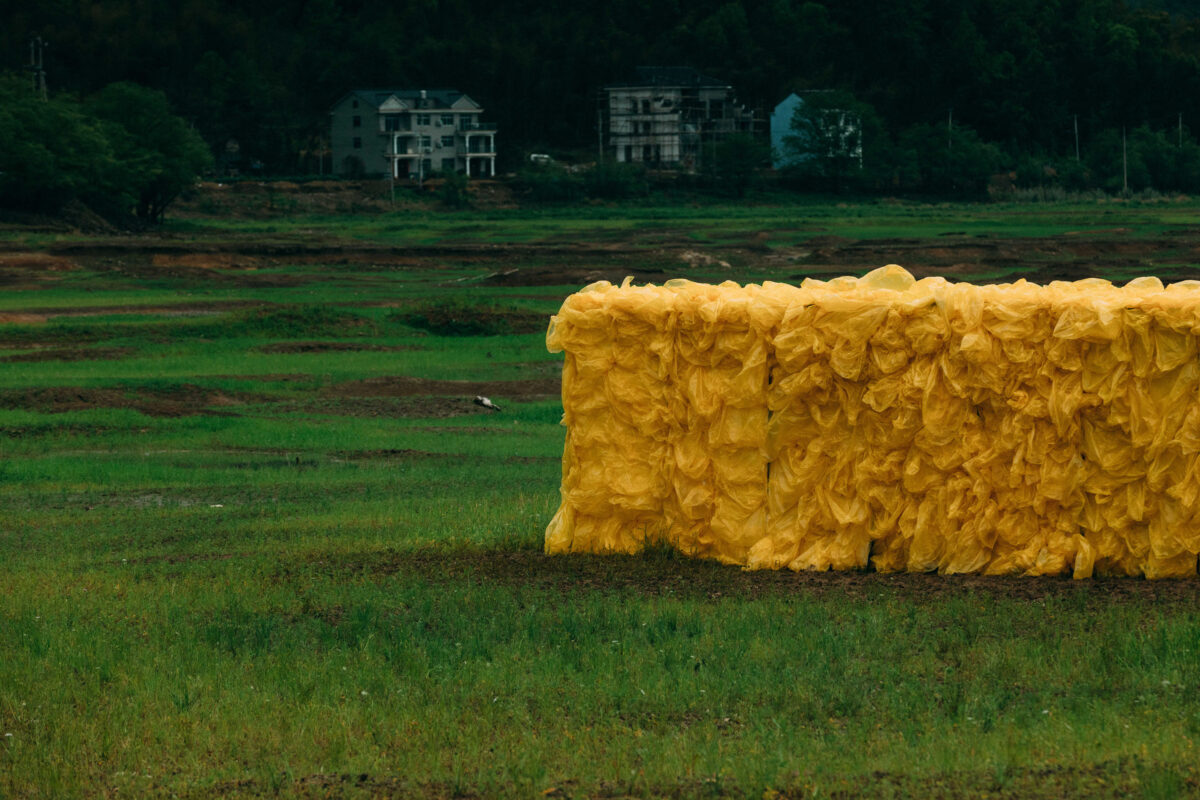 Video still of a large rectangular yellow sculpture of wood and plastic bags installed outdoors with houses
