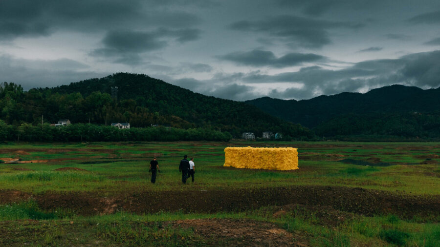 Video still of 3 people and a large rectangular yellow sculpture of wood and plastic bags installed outdoors