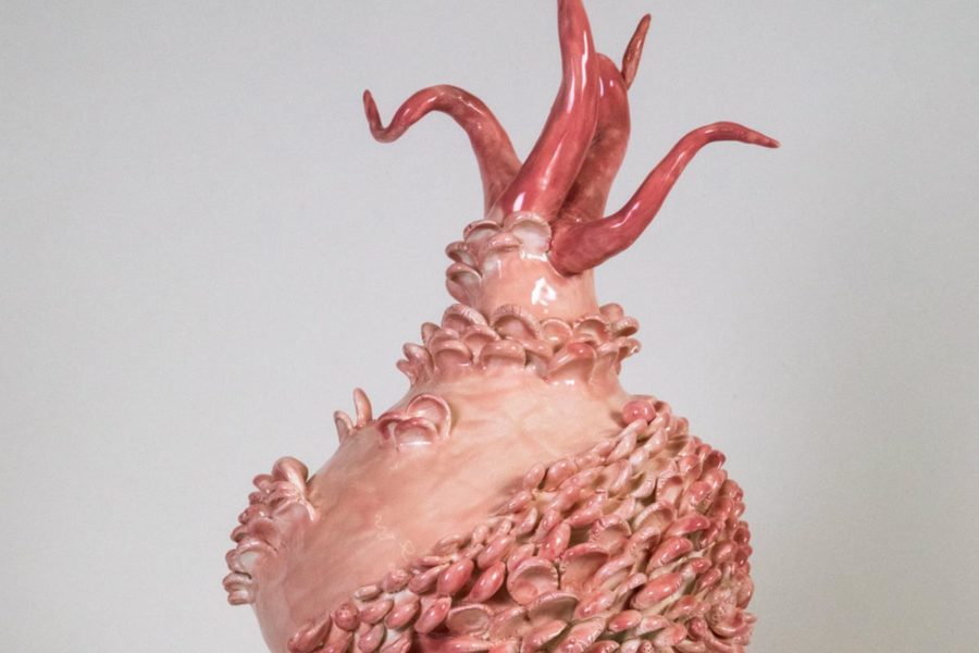 Ceramic Artwork by Carol Cao. The ceramic piece is colored pink. The overall shape is similar to a vase with a wide body with abstract shapes added onto the exterior. At the top are five forms that stick out from the top in a tentacle-like form. The background is solid grey.