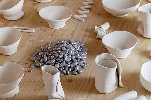 Ceramic sculptures of bowls and glasses are scattered around on the table, with a pile of silver beans in the middle of the image.