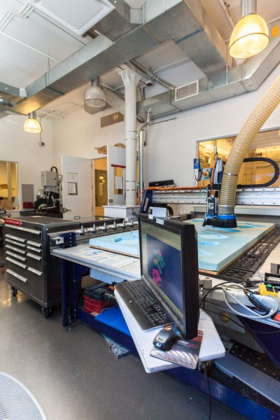 An interior view of the CNC Digital Sculpture lab. In the foreground is a large Shopbot CNC milling machine carving 3D shapes from a 4-foot by 8-foot sheet of blue polystyrene foam.