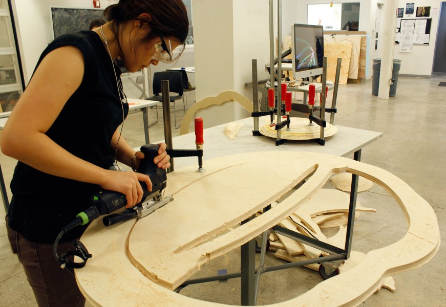 A student working in the main sculpture space, cutting an organic shape from a sheet of plywood using a Festool jigsaw.