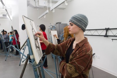 Students in a drawing class. In the center foreground, a student is standing at an easel making a charcoal drawing of a model that is not in the frame. In the background are more students standing at an easel and drawing.