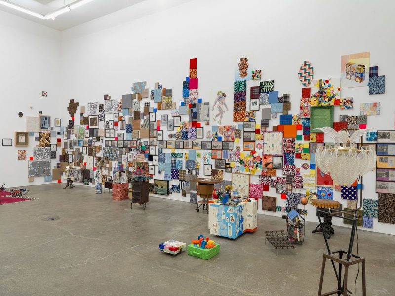 A photograph showing an installation by Nancy Shaver with many 2D works crowded together on the wall and various objects scattered around the floor.