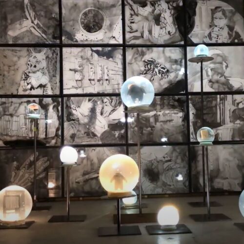16 ink drawings in lightboxes in front of them are large snow globes 33lbs each on steel stands at various heights. The room is dark and the objects are dramatically lit.