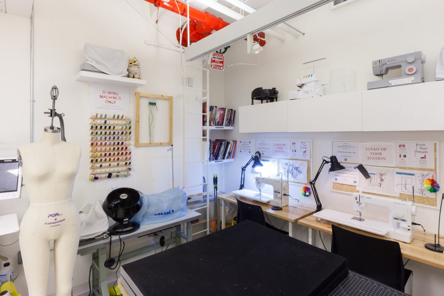 There is an overview of the fibers lab with a working table in the front. A female body-shaped mannequin, threads organized on the wall, sewing machines, and desk lamps.