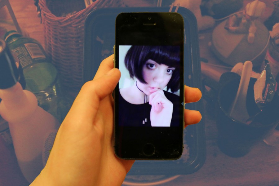 A portrait of a girl with short black hair, a hand on her lips, and she wears black. The image is on the phone held in hand, and in the background is a table full of bottles, glasses, and other objects