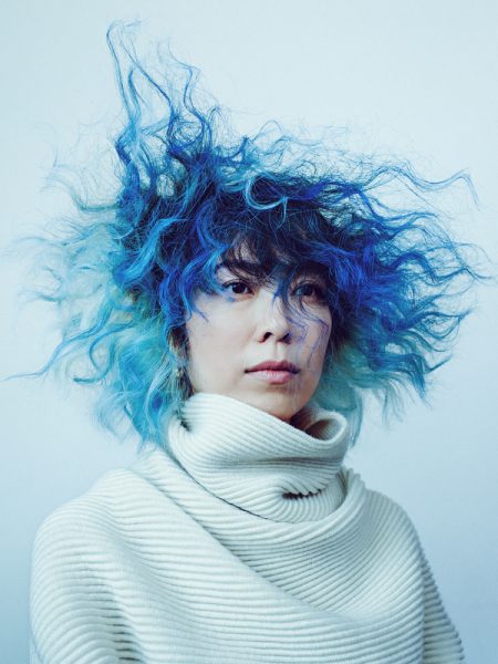 Portrait of Anicka Yi wearing a white ribbed sweater, with blue and black curly hair blowing in the wind, looking towards the right