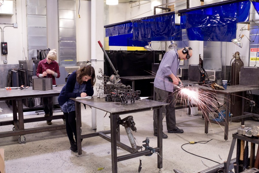 Students are working on different projects in the metal shop. On the left side in the background is a student working at a metal box, on the center of the image s a student taking notes in front of an assembled metal structure, and on the right side is a student cutting metal sheets with a grinder