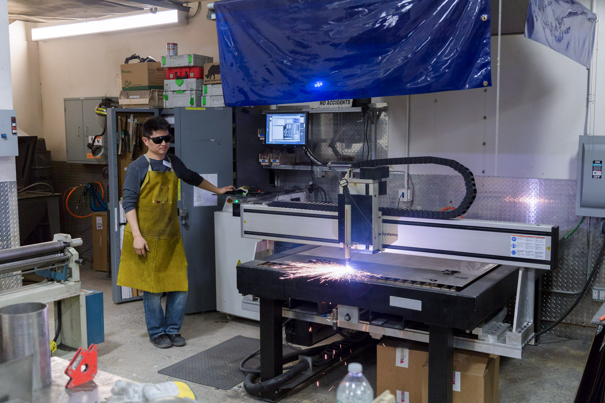 The student supervising the plasma cutting machine in the metal shop while cutting a sheet of metal after a provided design.