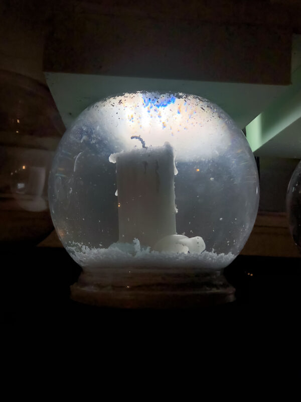 A small white candle sitting inside of a snow globe. The background is dark with a single light behind the globe.