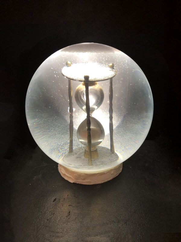A small hourglass sits inside of a glass snowglobe. The background is black.