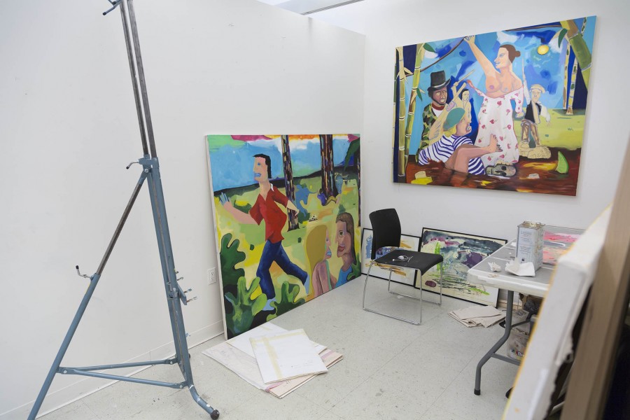 Installation view of an easel, a working table and a chair, a painting of two people about to kiss and a man with a red shirt running in the background, and another painting representing some people in the water with a woman wearing a white dress with flowers which have breasts uncovered by the men near her
