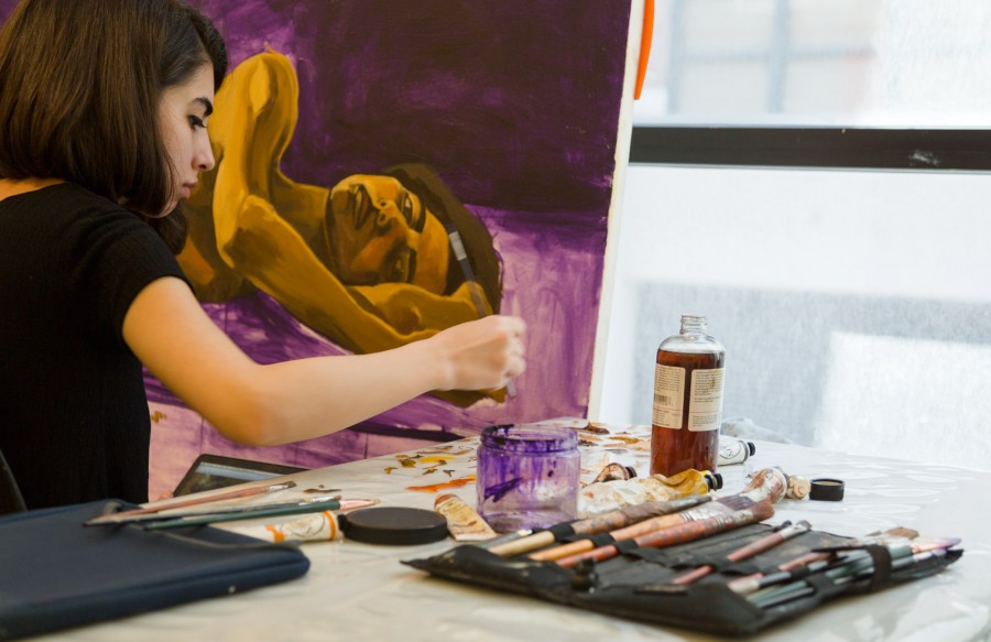 A student works on a painting of a woman laid down on a purple backdrop, with brushes, paints, and other materials on the table next to her