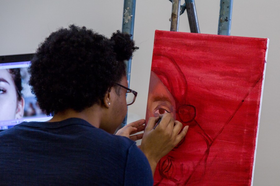 An SVA student is working on a portrait painting on a red background, and on her left side, she has a computer monitor with an image displayed.