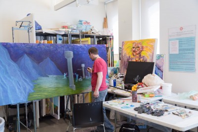 A student works on a large painting. The student is turned away from the camera and is in the middle of applying paint to a large painting that is in the center of the photograph. Behind the painting are large painting racks with paintings being stored for later use.