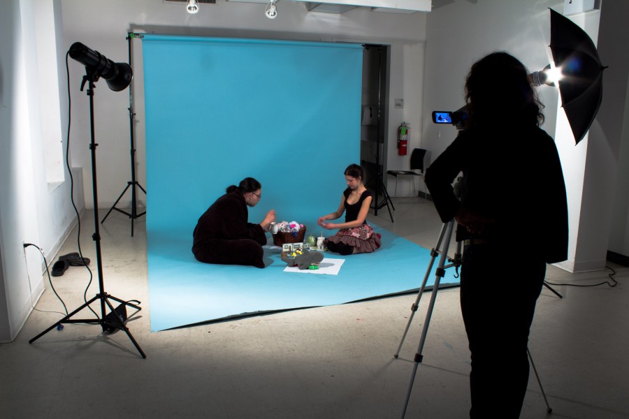 The photography class has one student handling the camera on a tripod and two other students sitting on a blue backdrop with props to create a scene.