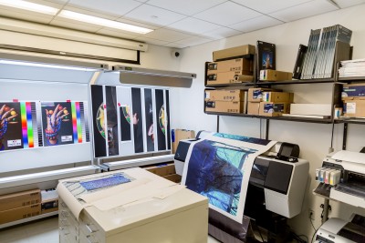Print room with several prints on the wall in the background and a big printer in the process of printing a large scale photograph