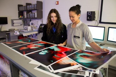 Two SVA students in the print room are looking at a bunch of large-scale prints on a table. 