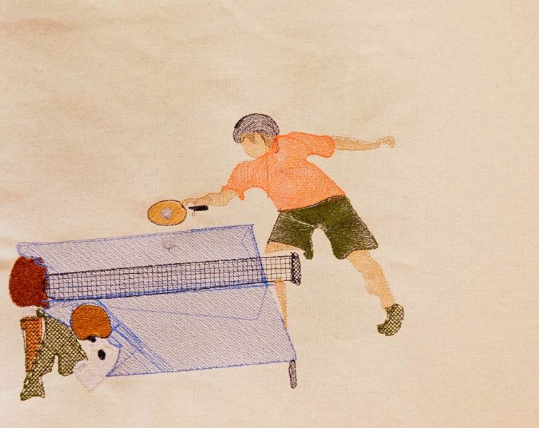 An embodiment with two people playing table tennis on a blue table. One person wears an orange T-shirt and green pants and has the ball, the other person is visible only with their rightsholder and wears a green t-shirt