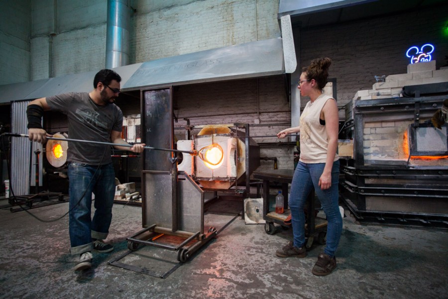 Two students in the glass and neon workshop, one is looking at the other, while he is modeling a bulb of glass in the oven