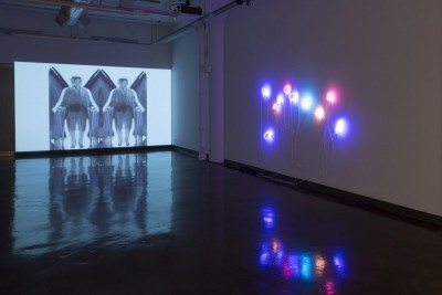 Installation view of an image projected on the wall of two human shapes with a triangular structure in their back and on the sidewall are installed small neon signs in blue, red, orange, and green