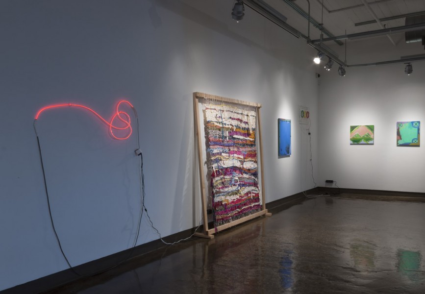 Installation view of a red neon sign, a piece of rug made from a lot of small pieces of multicolor fabrics, and paintings hung on the wall.