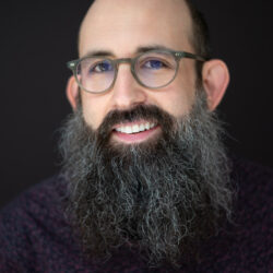 Photograph of Mike Falk. The photo is a headshot. Mike has a long beard and round glasses. He is smiling and looking at the camera. The background is black.