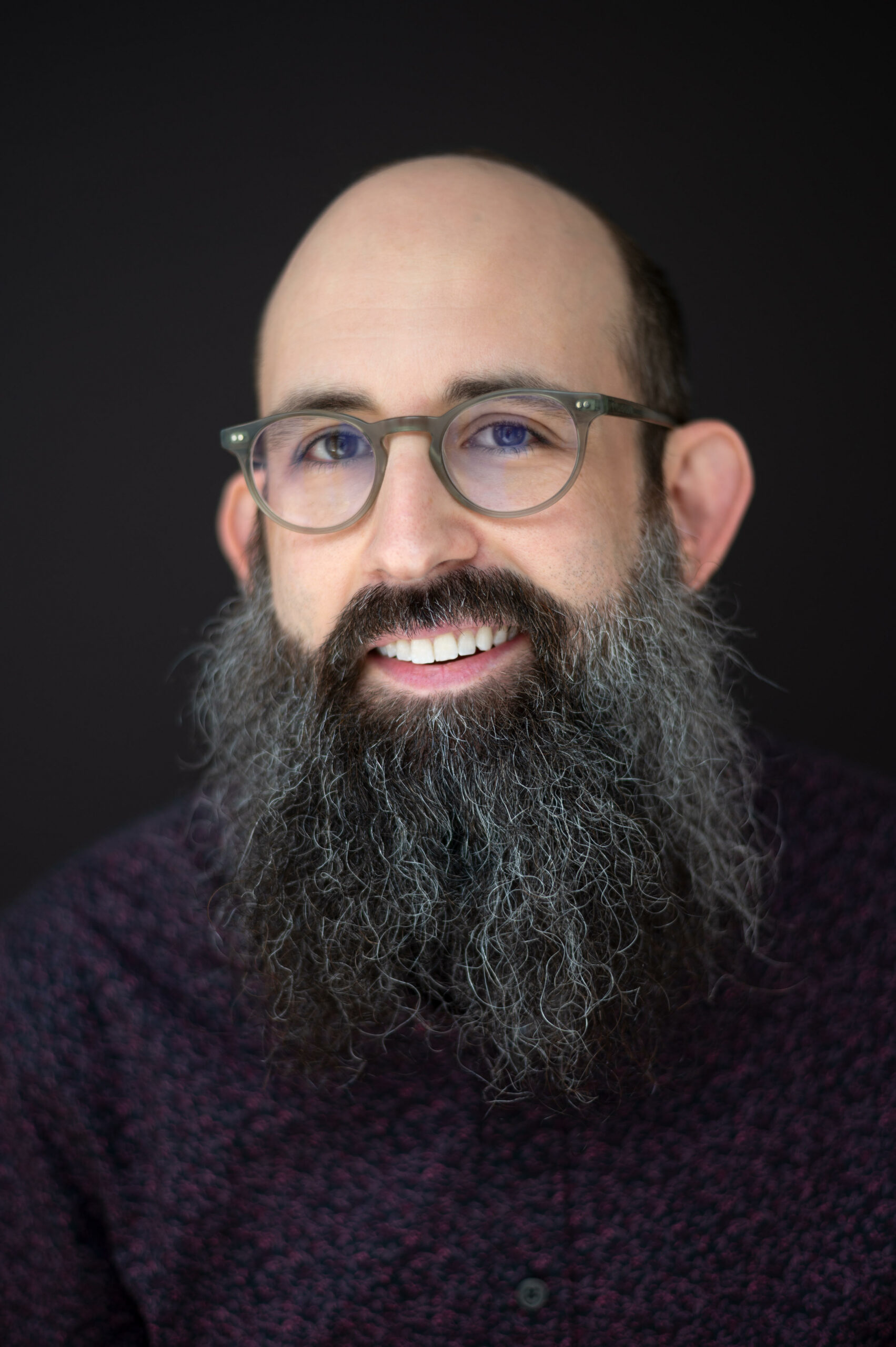 Photograph of Mike Falk. The photo is a headshot. Mike has a long beard and round glasses. He is smiling and looking at the camera. The background is black.