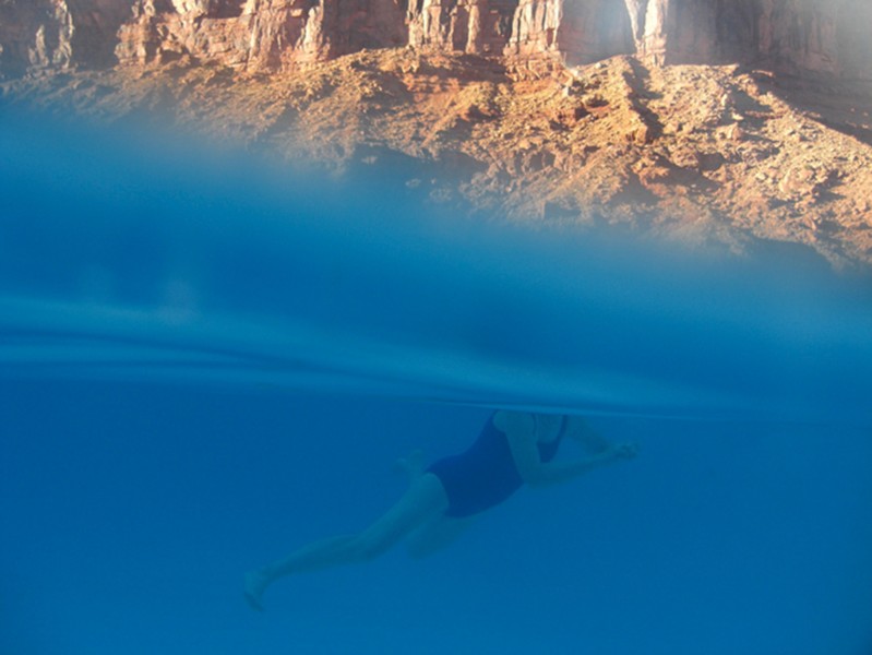 Person swimming in blue water and the top part of the frame shows an arid brown landscape.